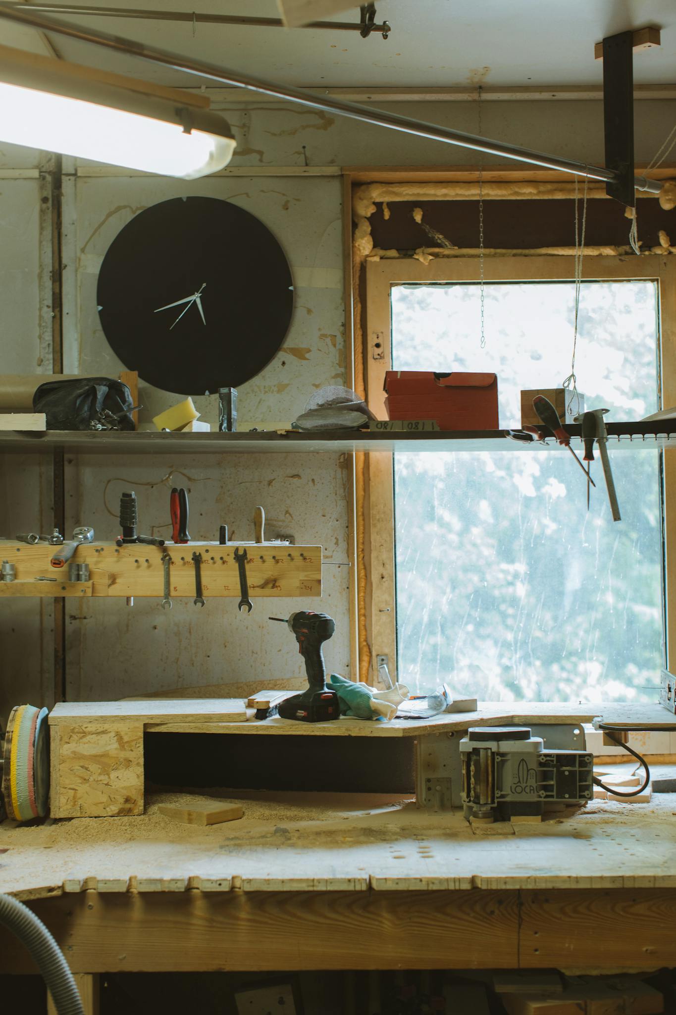 A rustic workshop with a variety of tools, a large clock, and a sunlit window.